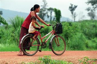 A mother in Lao holding a bike where a young girl is sitting in the back seat